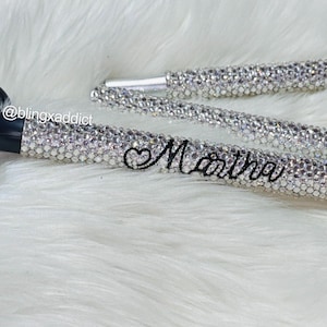 May include: A black mobility aid crutch featuring a handle and shaft covered in clear, sparkling rhinestones. The name Martha is written in black cursive script on the shaft, accompanied by a small heart symbol. The text @blingxaddict is visible.