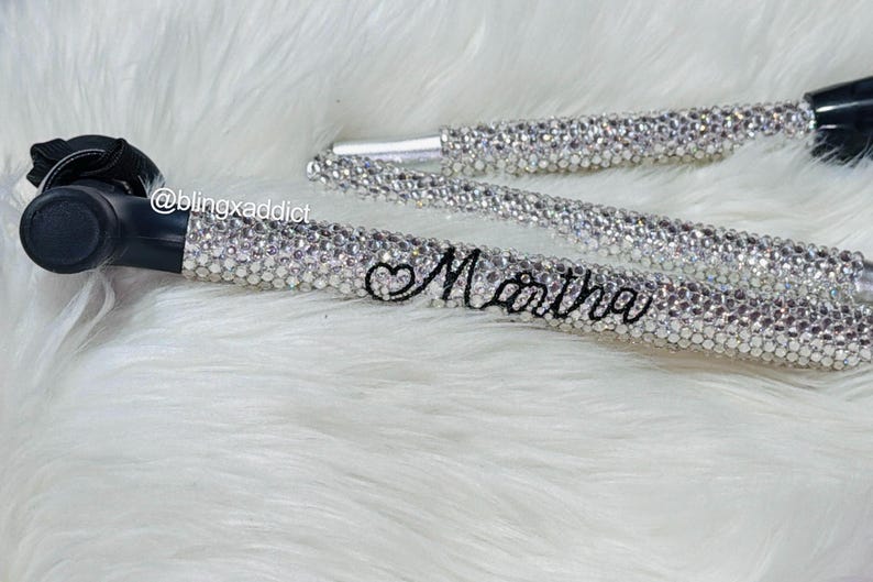 May include: A black mobility aid crutch featuring a handle and shaft covered in clear, sparkling rhinestones. The name Martha is written in black cursive script on the shaft, accompanied by a small heart symbol. The text @blingxaddict is visible.