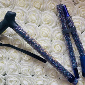 May include: A foldable walking cane featuring a black ergonomic handle and a shaft covered in iridescent blue and purple rhinestones. The cane includes a black wrist strap and a blue rubber tip. The text @blingxaddict is visible on the shaft.