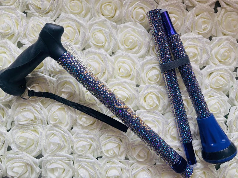 May include: A foldable walking cane featuring a black ergonomic handle and a shaft covered in iridescent blue and purple rhinestones. The cane includes a black wrist strap and a blue rubber tip. The text @blingxaddict is visible on the shaft.