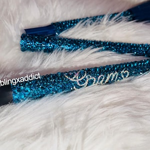 May include: A foldable walking cane covered in shimmering teal rhinestones. The handle is black with a black strap. The text @blingxaddict is printed in white near the handle, and the word Grams is spelled out in clear crystals on the shaft.