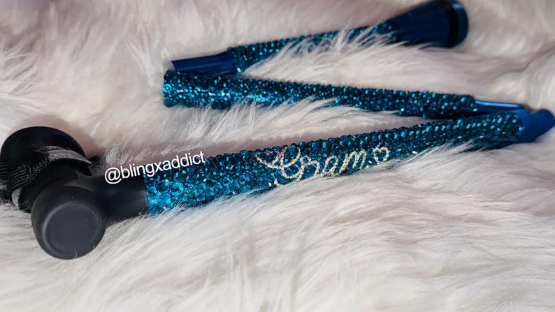 May include: A foldable walking cane covered in shimmering teal rhinestones. The handle is black with a black strap. The text @blingxaddict is printed in white near the handle, and the word Grams is spelled out in clear crystals on the shaft.