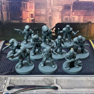 May include: A collection of ten unpainted gray plastic miniature figures standing on circular bases. Each figure depicts a character in tactical gear, some holding small weapons or equipment, arranged in a group formation on a textured surface.