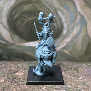 May include: A grey plastic miniature figure of a goblin-like creature wearing a pointed hat, holding a sword and a small round shield, mounted on a large spherical creature with a wide mouth and sharp teeth, standing on a square black base.