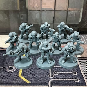 May include: A collection of nine unpainted gray plastic miniature soldier figurines standing on circular bases. Each figure is depicted in a combat stance, wearing tactical gear and helmets, and holding various rifles or automatic weapons.
