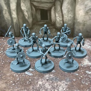 Könnte beinhalten: Eine Sammlung von zehn unbemalten Skelett-Miniaturen aus grauem Kunststoff auf runden Sockeln. Jede Figur ist in einer dynamischen Pose dargestellt, hält ein Schwert in der einen und einen runden Schild in der anderen Hand und zeigt Details.