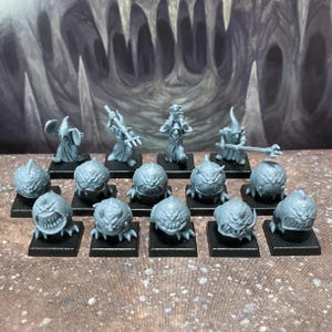 May include: A collection of fourteen unpainted grey plastic miniature figures on black square bases. The group includes four hooded humanoid figures holding various weapons and ten round, toothy, multi-eyed creatures with small claws.