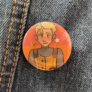 May include: A round button pin featuring an illustration of a character with blonde hair and a yellow outfit. The background is a gradient of orange and pink with star accents. The pin is set against a denim background.