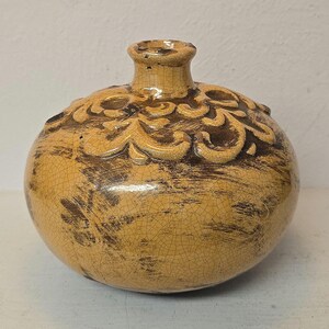 May include: A round ceramic vase with a narrow neck and a wide, bulbous body. The surface features a glossy, mustard-yellow glaze with a dark brown crackle pattern and a raised, embossed scrollwork design around the top shoulder area.