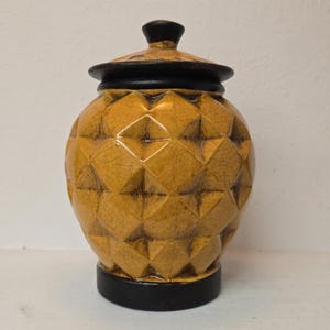 May include: A ceramic jar with a lid, featuring a mustard yellow body with a raised diamond-shaped geometric pattern and a fine crackle glaze finish. The jar has a solid black circular base and a matching black lid with a small central knob.