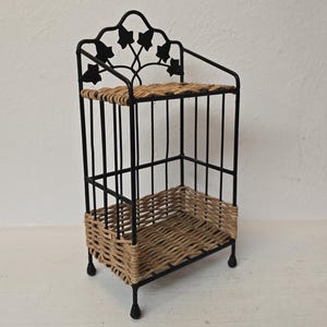 May include: A two-tier shelf unit featuring a black metal frame with four thin legs and a decorative top arch adorned with metal ivy leaf cutouts. The two shelves are constructed from woven light brown wicker, providing a natural contrast.