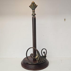 Puede incluir: Un soporte vertical para papel de cocina con una base circular de madera marrón oscuro y un poste central. La parte superior del poste tiene un remate de flor de lis en tono dorado. Un adorno de metal curvado en acabado bronce oscuro descansa sobre la base.