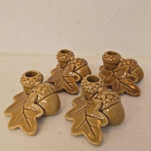 Puede incluir: Conjunto de cinco candelabros de cerámica con forma de bellotas y hojas de roble. Los candelabros son de color marrón cálido. Cada uno está diseñado para sostener una vela pequeña, ofreciendo un elemento decorativo natural.
