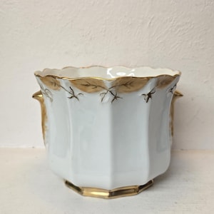 Puede incluir: Jardinera de porcelana blanca con borde dorado y diseño de hojas. La jardinera tiene una forma geométrica con un borde festoneado y asas pequeñas. Los detalles dorados contrastan con la porcelana blanca.