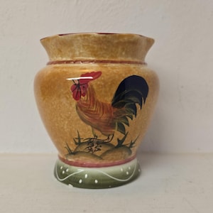 Puede incluir: Un jarrón de cerámica con un diseño de gallo. El jarrón tiene un fondo marrón con un gallo pintado en tonos rojos, naranjas y marrones. La base es verde con puntos blancos y el borde es rojo.