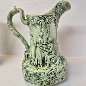 Op de afbeelding: Een groene keramische kan met een reliëfdesign van een stel dat in een tropische scène met palmbomen staat. De kan heeft een brede opening en een handvat dat omhoog en over de bovenkant buigt.