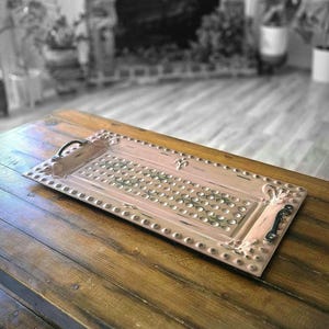 May include: A rectangular, distressed, blush-colored metal tray with a dotted center design and decorative edges. The tray has two black handles and is set on a wooden table. The tray is approximately 20 inches long.