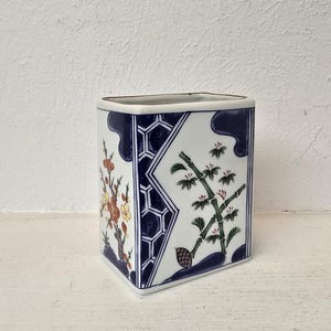 Puede incluir: Un jarrón cuadrado de cerámica con fondo blanco y ribete azul. El jarrón presenta diseños florales y de bambú en tonos rojos, amarillos y verdes. El jarrón mide aproximadamente 13 cm de alto.