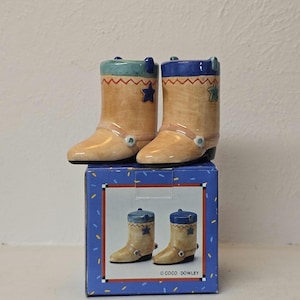 Könnte beinhalten: Zwei Cowboy-Stiefel-Figuren aus Keramik in Beige, Blau und Türkis. Jeder Stiefel hat einen Stern und einen dekorativen Riemen. Die Stiefel stehen auf einer blauen Schachtel mit einem passenden Bild. Auf der Schachtel steht "COCO DOWLEY".