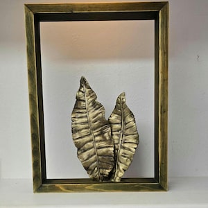 Könnte beinhalten: Dekorative Wandkunst mit zwei strukturierten, goldfarbenen Blätterskulpturen in einem Holzrahmen. Der Rahmen hat eine dunkle, abgenutzte Oberfläche, und die Blätter haben detaillierte Adern.