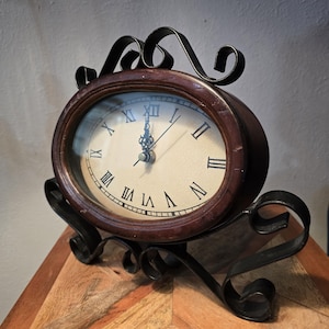 May include: A brown and black clock with Roman numerals on a white face. The clock is on a black metal stand with a decorative scroll design. The clock is sitting on a wooden surface.