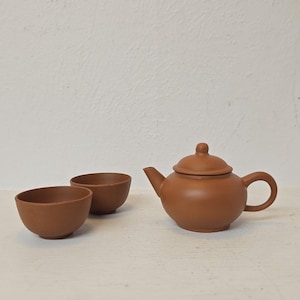 Könnte beinhalten: Eine braune Keramik-Teekanne mit passendem Deckel und zwei kleinen Schalen. Die Teekanne hat einen abgerundeten Körper, einen gebogenen Ausguss und einen Schlaufengriff. Die Schalen sind rund und stehen neben der Teekanne. Das Set steht auf einer weißen Oberfläche.
