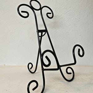 May include: Black metal display stand with decorative scrollwork. The stand has a three-legged base and a back support with a curved top. The stand is designed to hold a picture frame or small decorative item.