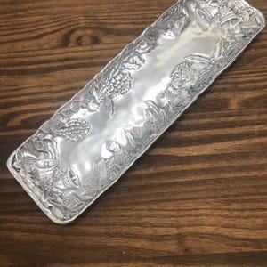 May include: A silver serving tray with a raised design of grapes and leaves. The tray is rectangular and has a slightly curved edge.