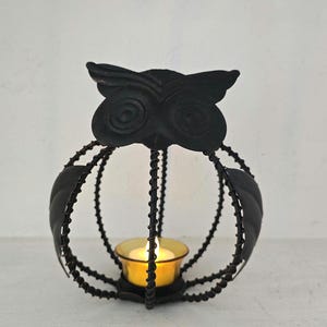 May include: Black metal owl-shaped candle holder with a lit yellow candle inside. The owl has a decorative cage design with leaf accents. The candle holder sits on a white surface.