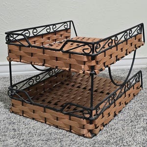 Könnte beinhalten: Zweistöckiger Schreibtisch-Organizer mit einem gewebten, hellbraunen Korbdesign. Der Organizer hat einen schwarzen Metallrahmen mit dekorativen Schnörkeln. Die beiden Ablagen sind gestapelt und bieten viel Platz zum Ordnen von Papieren und Bürobedarf.