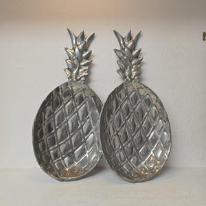 May include: Two silver-coloured metal trays shaped like pineapples. Each tray features a textured, diamond-patterned body and a detailed, pointed leaf top. The metallic surface has a polished finish with visible indentations and contours.