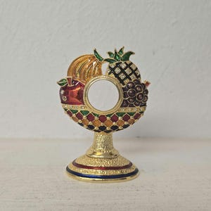 May include: A gold-colored decorative stand with a circular frame featuring enamel fruit designs, including a banana, apple, pineapple, and grapes. The frame has a central opening and is adorned with colorful geometric patterns and small crystals.