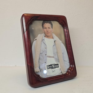 May include: A dark reddish-brown wooden picture frame with a glossy finish and a scalloped inner edge. The frame contains a display photo of a person wearing a light-coloured jacket and sweater, with a label reading Rare Woods 5x7 visible on the glass.