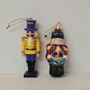 May include: Two Christmas ornaments in the shape of nutcrackers. One is a soldier in a yellow jacket and blue pants. The other is a drummer in a red jacket. Both have gold string hangers.