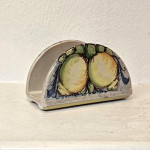 May include: A ceramic napkin holder with a semi-circular design. The holder is painted with a design of two yellow fruits with green leaves and blue accents. The base is a light beige color.