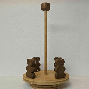 Może przedstawiać: Drewniana karuzela z centralnym słupem i okrągłą podstawą. Do podstawy przymocowane są cztery małe, brązowe figurki misiów. Karuzela wykonana jest z jasnobrązowego drewna i stoi na białej powierzchni.