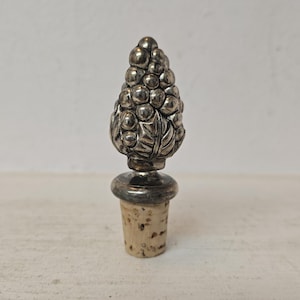 May include: A silver-toned bottle stopper featuring a decorative top shaped like a cluster of grapes with detailed leaves. The metallic topper is mounted on a tapered, light brown natural cork base, designed for sealing bottles securely.