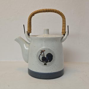 Könnte beinhalten: Weiße Keramik-Teekanne mit einer Hahn-Illustration, einem geflochtenen Griff und einem dunkelblauen Sockel. Die Teekanne hat einen Ausguss und einen Deckel. Der Hahn ist in Blau und Schwarz dargestellt. Die Teekanne ist mit kleinen schwarzen Punkten gesprenkelt.