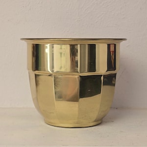 Puede incluir: Una maceta dorada brillante con un diseño geométrico. La maceta tiene una forma ancha y redondeada con un borde ligeramente ensanchado. La superficie refleja la luz, creando una apariencia metálica brillante. Esta maceta decorativa es adecuada para uso en interiores o exteriores.