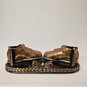 May include: A pair of gold-toned metal shoes with a bow on the base. The shoes are on a decorative base with a rope-like design.