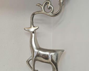 May include: A silver-colored deer figurine with a candle holder. The deer has a slender body, a long neck, and a decorative antler design. The candle holder is a small, round cup attached to the deer's antler. The figurine is made of metal.
