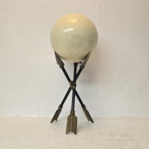May include: A decorative sphere in a light green hue rests on a black metal stand. The stand features three arrows with brass-colored arrowheads, creating a unique and artistic display. The sphere is the focal point of this decorative piece.
