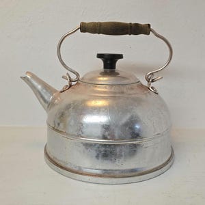 Puede incluir: Una tetera plateada vintage con un pico, un pomo negro y un asa de madera. La tetera tiene un cuerpo redondeado y un asa de metal curvada. El asa es de madera y metal. La tetera está sobre una superficie blanca.