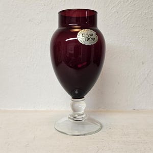 May include: A ruby-colored glass vase with a bulbous body and a clear, stemmed base. The vase has a label that reads "Royal Ruby". The vase is likely used for decorative purposes or displaying flowers.
