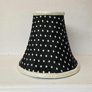 May include: A black lampshade with white polka dots. The shade has a classic bell shape and is trimmed with a cream-colored ribbon along the top and bottom edges. The lampshade is designed to fit a small lamp base.
