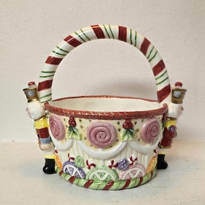 Puede incluir: Una cesta de cerámica con asa y borde a rayas de bastón de caramelo. La cesta está decorada con adornos coloridos, espirales y figuras de cascanueces a cada lado. La cesta mide aproximadamente 20 cm de altura.