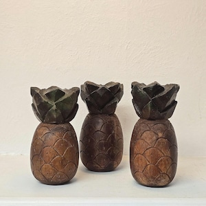 May include: Three wooden pineapple sculptures with detailed carved textures. The pineapples have a dark brown color with a lighter brown base. The tops are a darker shade of brown. The sculptures are displayed on a white shelf.