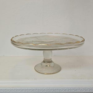 May include: A clear glass cake stand with a scalloped edge and gold trim. The stand has a round base and a decorative stem. The cake stand is perfect for displaying desserts.