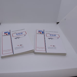 Pode incluir: Dois manuais do proprietário do avião Cessna 152 brancos com detalhes em vermelho e azul. Os manuais são intitulados "Manual de Operação do Piloto" e "Manual do Proprietário".