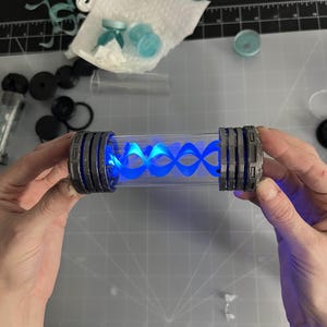 May include: A clear, cylindrical container with gray and silver accents on each end. The container holds a glowing blue, spiral-shaped light. The item is held by two hands against a gray cutting mat.